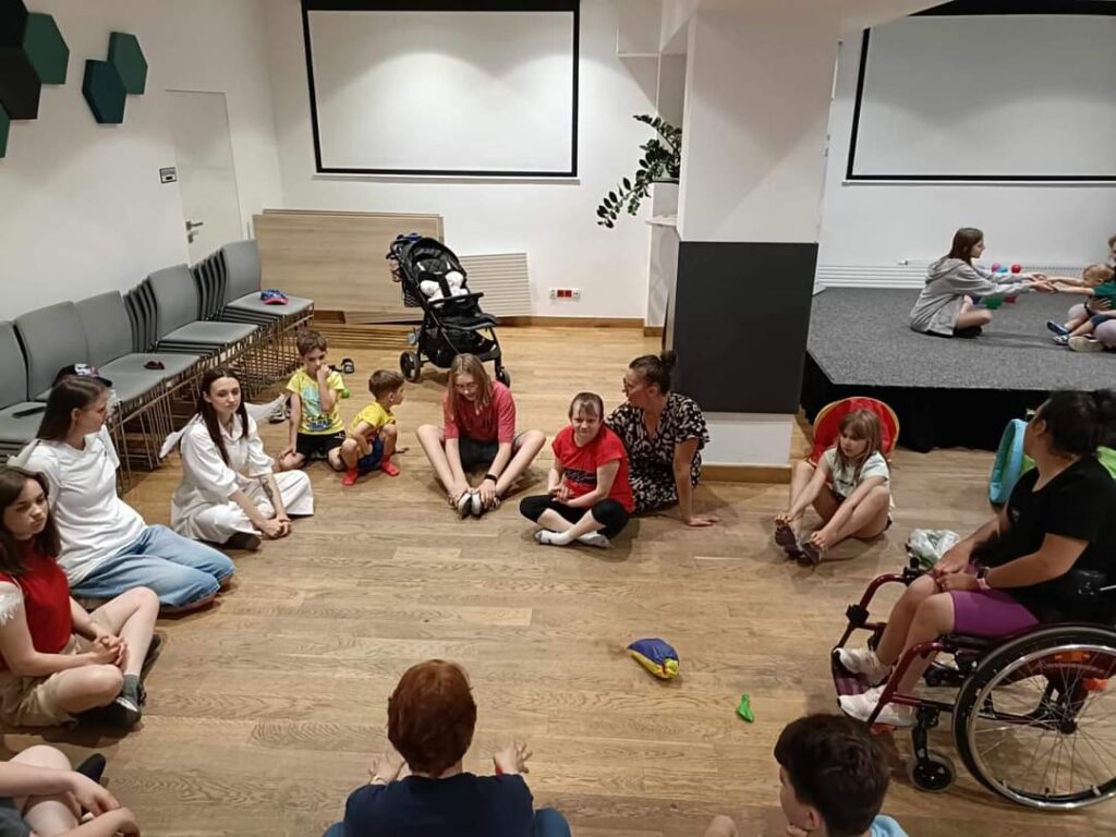 dzieci i wolontariusze siedzący na podłodze i dziewczyna na wózku